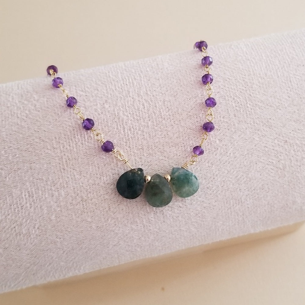 A beaded necklace with purple amethyst beads, gold spacers, and three green teardrop gemstones at the center, displayed on a textured white jewelry stand.
