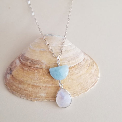 A silver necklace with a pendant featuring a light blue aquamarine half moon stone and a teardrop-shaped iridescent moonstone stone, displayed on a beige clam shell.