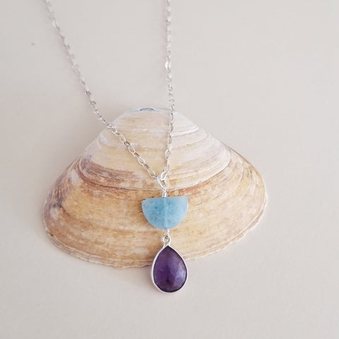 A silver necklace with a unique pendant featuring a light blue aquamarine semicircle stone and a dangling purple amethyst teardrop stone, displayed on a natural clam shell against a neutral beige background.