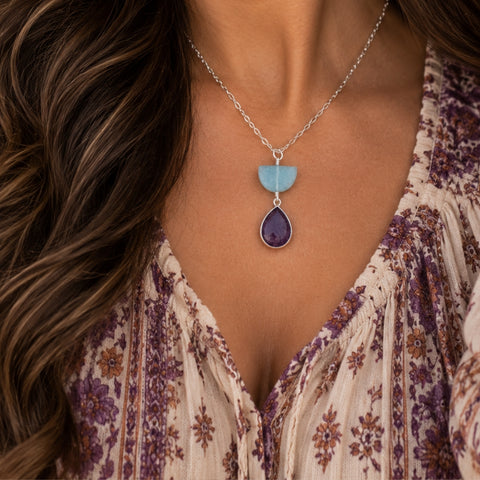 A close-up of a woman wearing a silver pendant necklace with a two-stone design: a light blue teardrop-shaped stone on top of a larger purple teardrop-shaped stone, paired with a cream floral blouse.