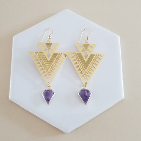 Gold triangle earrings with purple gemstones on a white hexagonal stand.