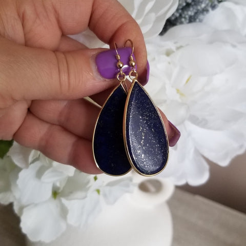 Teardrop-shaped earrings with gold accents held by a hand against a white flower background. Makes a great gift for her