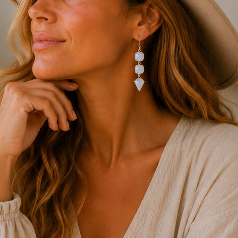 A close-up lifestyle shot of a woman wearing bohemian-style dangle earrings made of natural moonstone and gold-toned metal, paired with a beige hat and a light beige top.