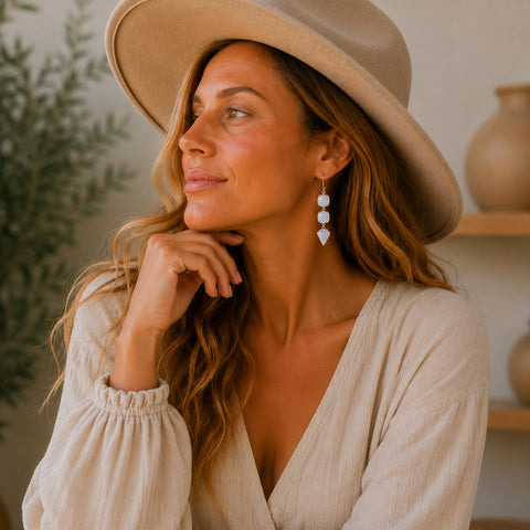 A woman wearing a beige wide-brim hat and a cream-colored top, looking thoughtfully to the side while wearing geometric moonstone dangle earrings.