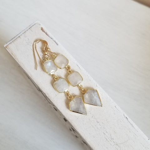A pair of gold drop earrings with irregular milky white moonstones, displayed on a distressed white wooden block against a light neutral background.
