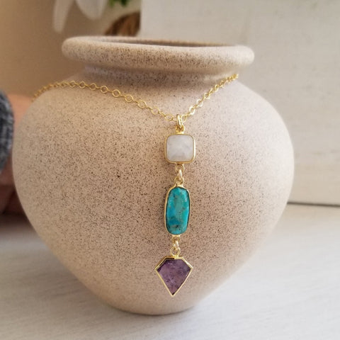 A gold chain necklace with three dangling gemstone charms, displayed on a textured beige ceramic vase. The charms are a square white stone, an oval turquoise blue stone, and a diamond-shaped purple stone.